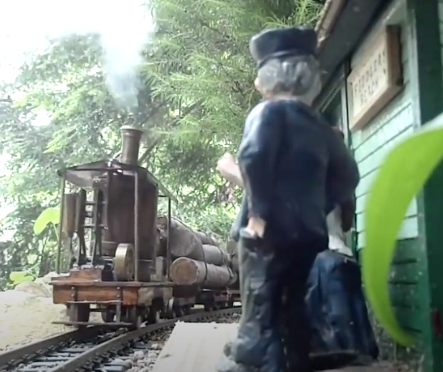 Homemade garden-scale live steam logging locomotive passes near a figure posed at a train depot.