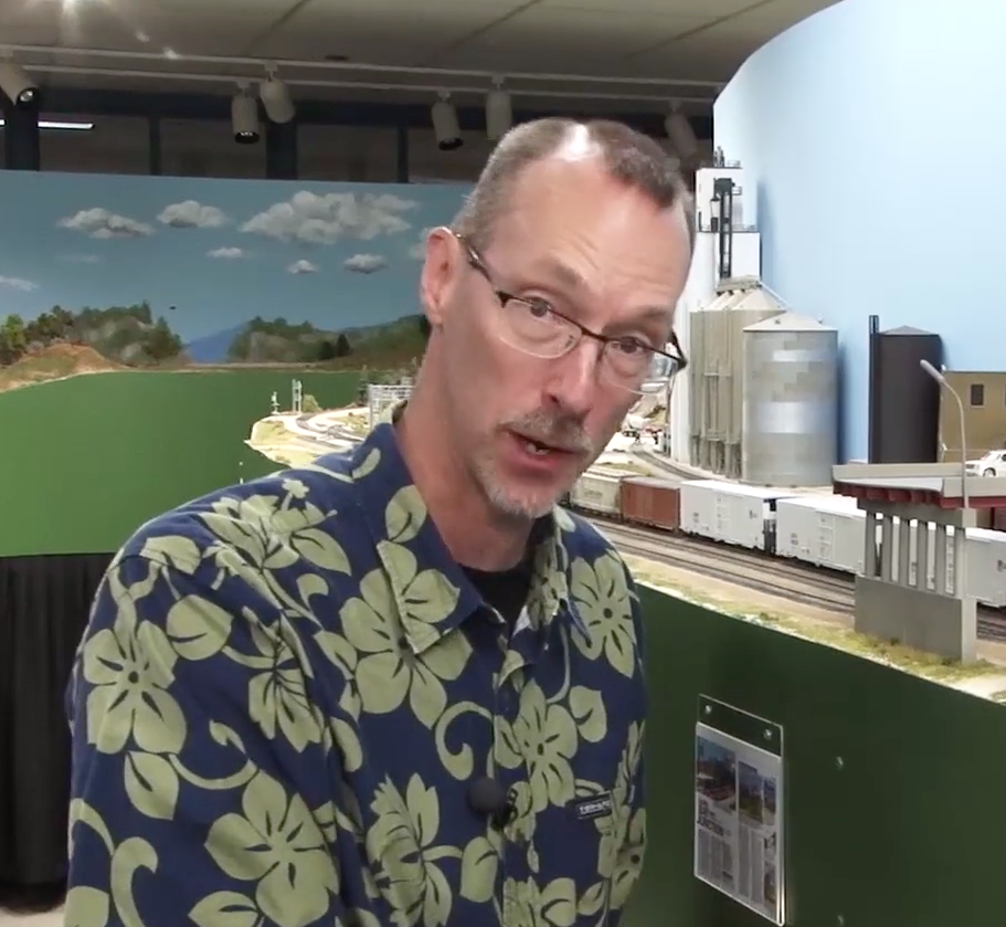 Man wearing a Hawaiian-style shirt facing camera before a model train layout.
