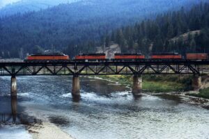 Orange and black diesel and electic locomotives on deck truss bridge over river