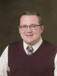 A headshot of a man in a brown sweater vest with a plaid tie and glasses smiling in front of a brown photo background