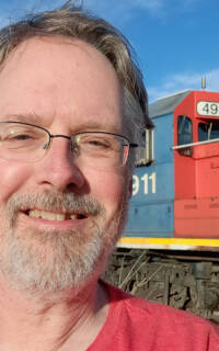A man in a red t shirt stands in front of a red and blue locomotive