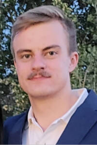 A man in a navy blue suit with a white shirt with a green foliage background