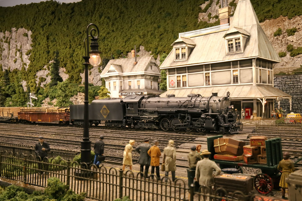 A 2-8-4 steam locomotive model pulls a train through a center of a town depicted in a mountainous, forested landscape.