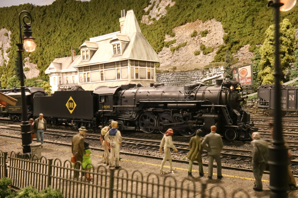 A 4-6-2 steam locomotive model pulls a train through the center of a town in a mountainous and forested scene.