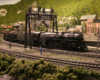 A 4-8-4 steam locomotive leads a freight train through an S-curve in a mountainous and forested scene and past several railroad and industrial buildings.