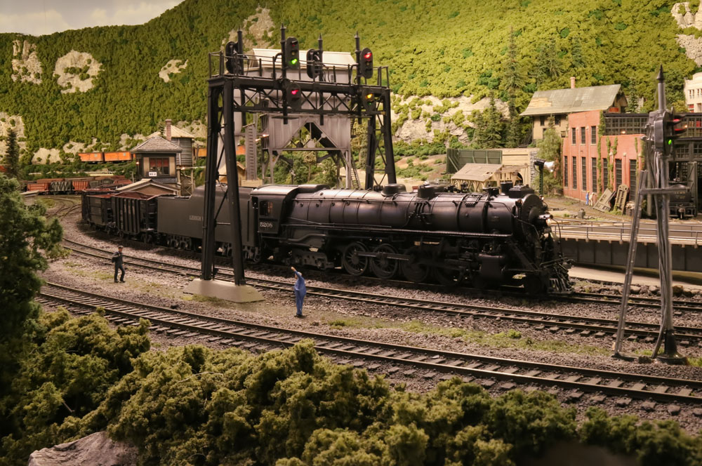 A 4-8-4 steam locomotive leads a freight train through an S-curve in a mountainous and forested scene and past several railroad and industrial buildings.
