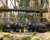 Three model steam locomotives pause in an industrial scene surrounded by trees.