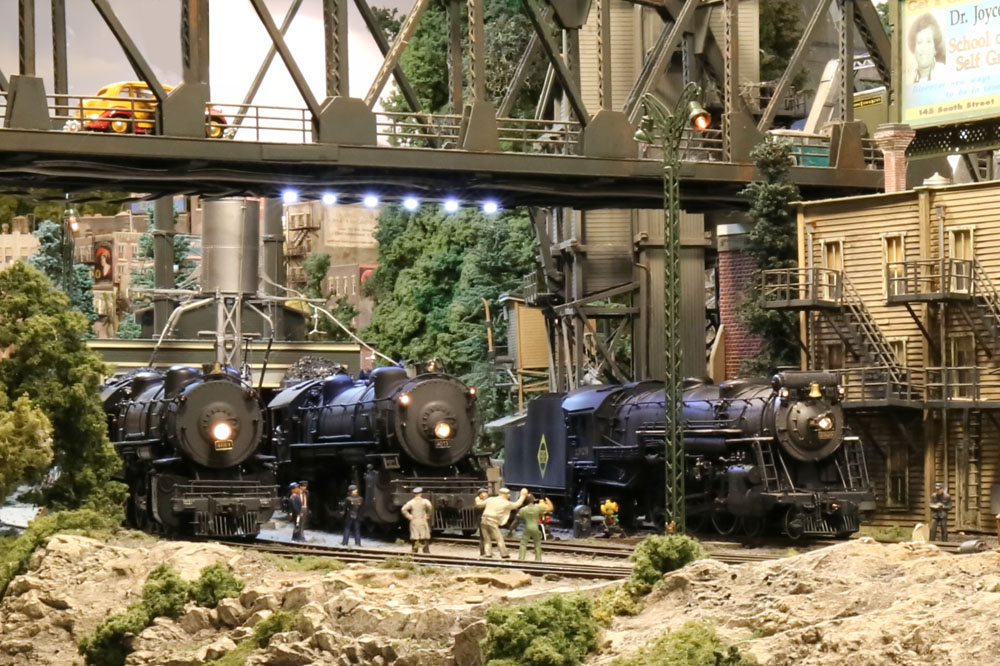 Three model steam locomotives pause in an industrial scene surrounded by trees.