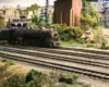 A 2-10-2 model steam locomotives on yard tracks amid a verdant industrial scene.