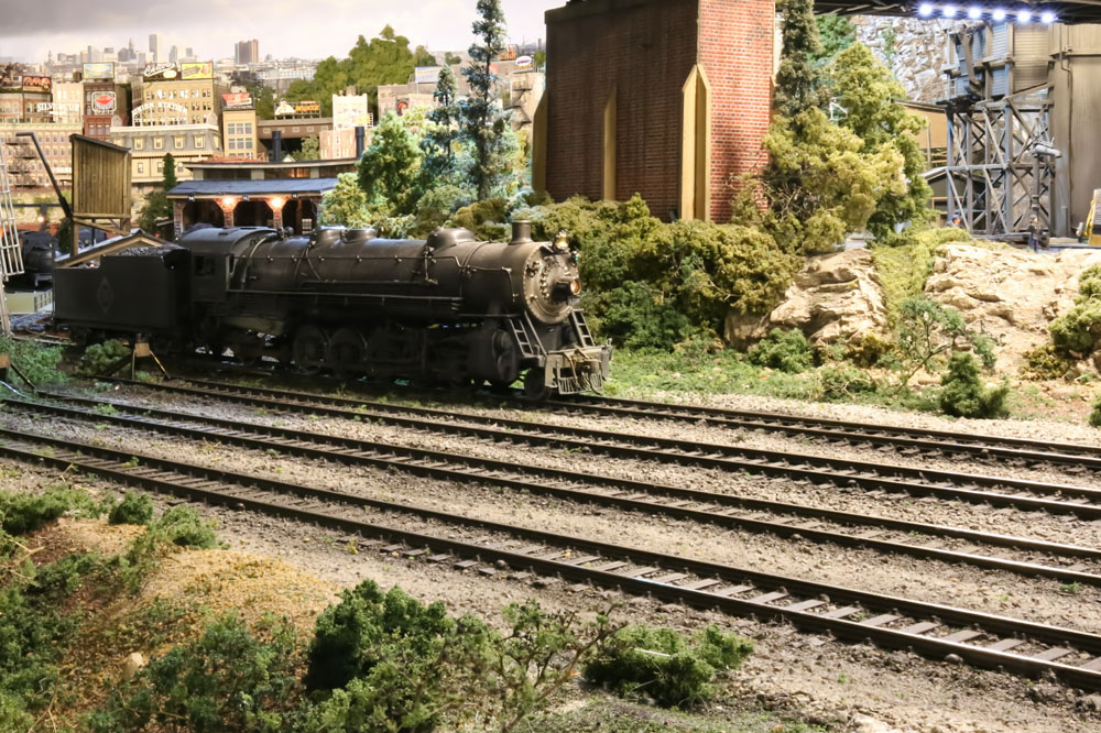 A 2-10-2 model steam locomotives on yard tracks amid a verdant industrial scene.