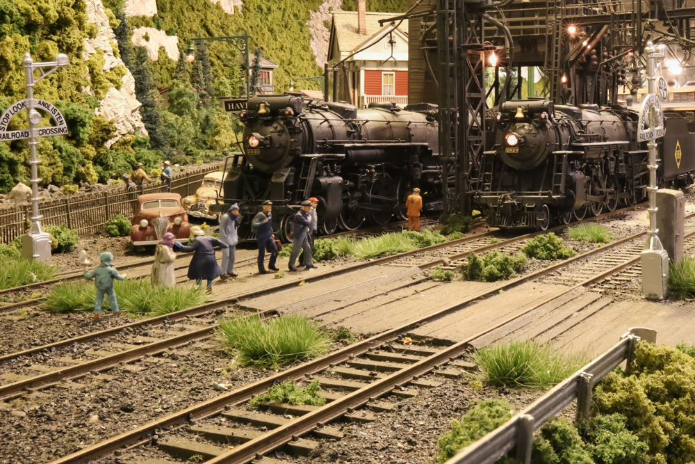 Model people cross tracks at a crossing with two model steam locomotives just behind them to the right.