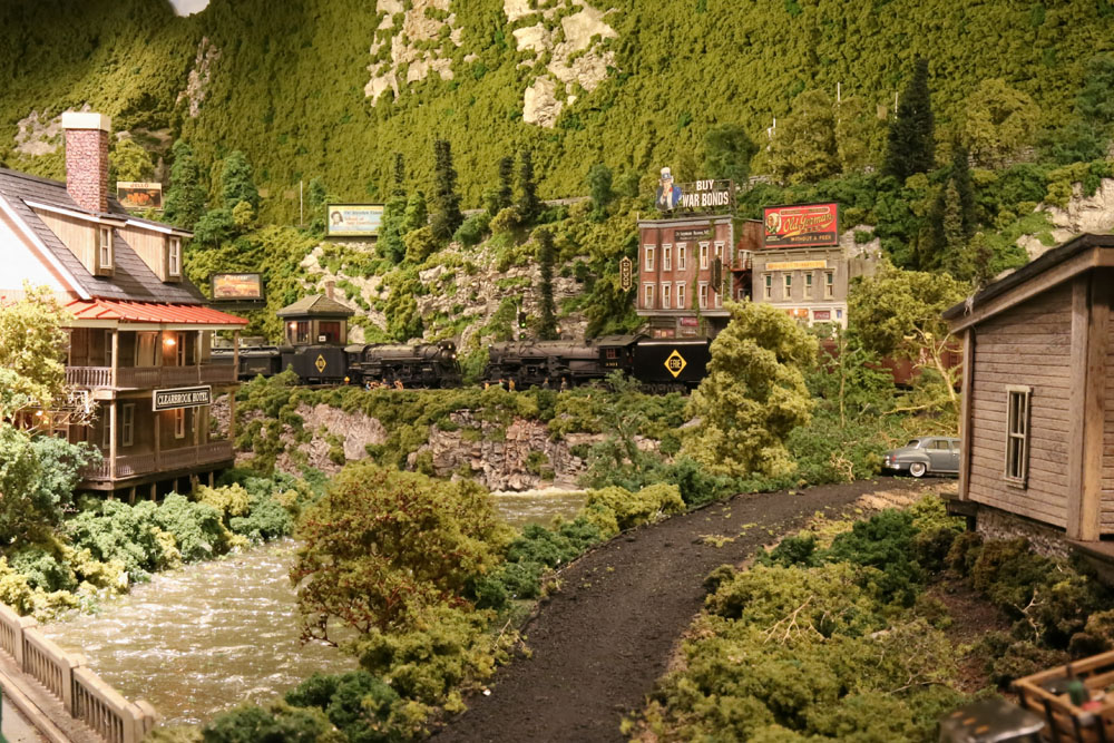 Two steam locomotives powered trains meet in a mountainous, forested landscape bracketed by rustic late-19th century wood or brick buildings. Appalachian.