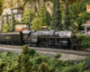 A 2-8-4 steam locomotive runs through a verdant, rocky scene.