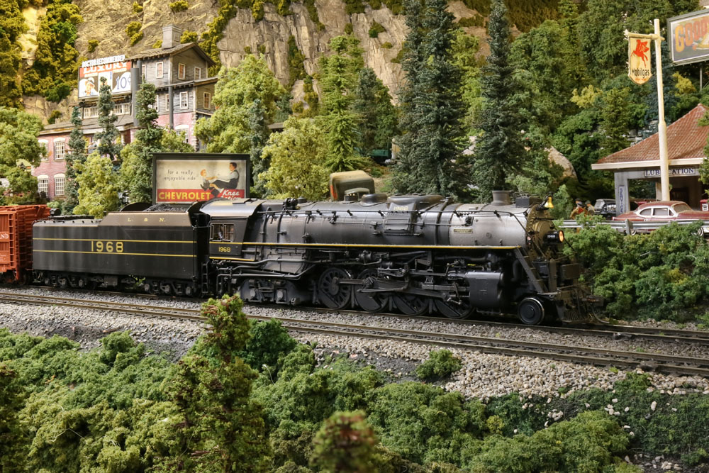 A 2-8-4 steam locomotive runs through a verdant, rocky scene.