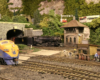 A model steam locomotive exits a tunnel into a rail yard. A blue and yellow diesel locomotive sits in the foreground.