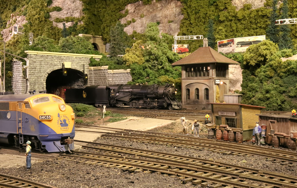 A model steam locomotive exits a tunnel into a rail yard. A blue and yellow diesel locomotive sits in the foreground.