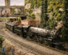 A 4-6-2 model steam locomotive pulls a passenger train from a yard around a curve in a verdant town scene. A rail yard with road bridge, signals, and additional steam trains are in the middle and background.