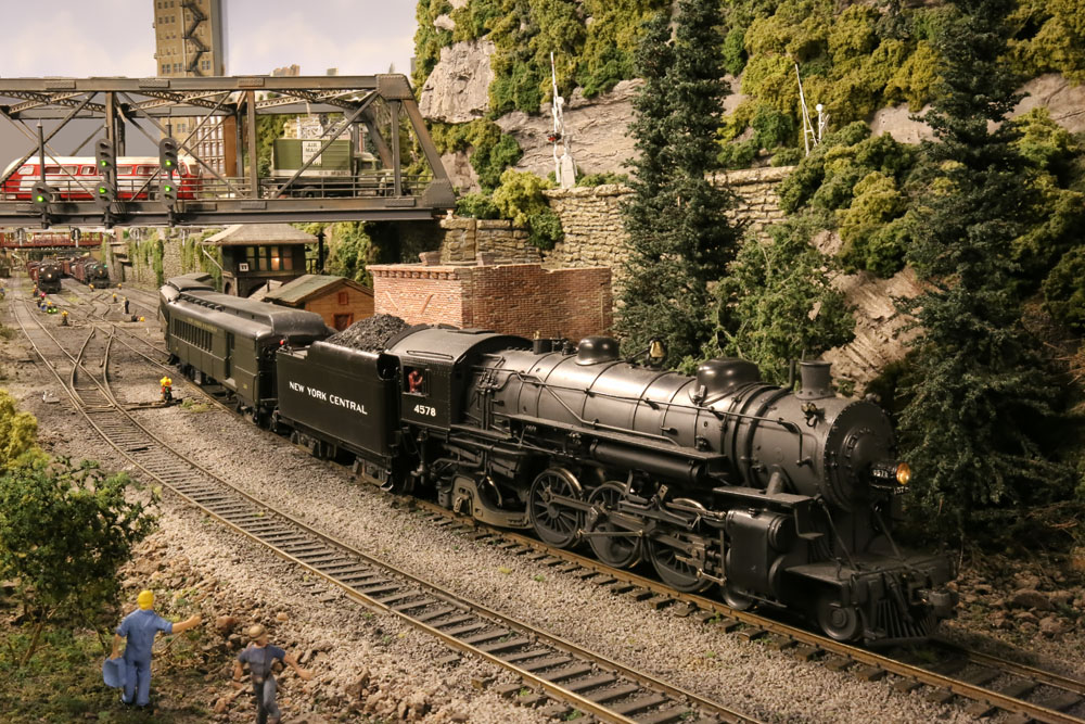 A 4-6-2 model steam locomotive pulls a passenger train from a yard around a curve in a verdant town scene. A rail yard with road bridge, signals, and additional steam trains are in the middle and background.