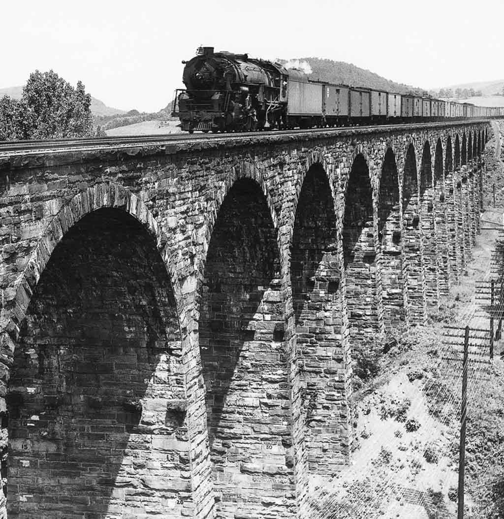 Black-and-white photo of steam-powered freight train crossing stone-built viaduct.