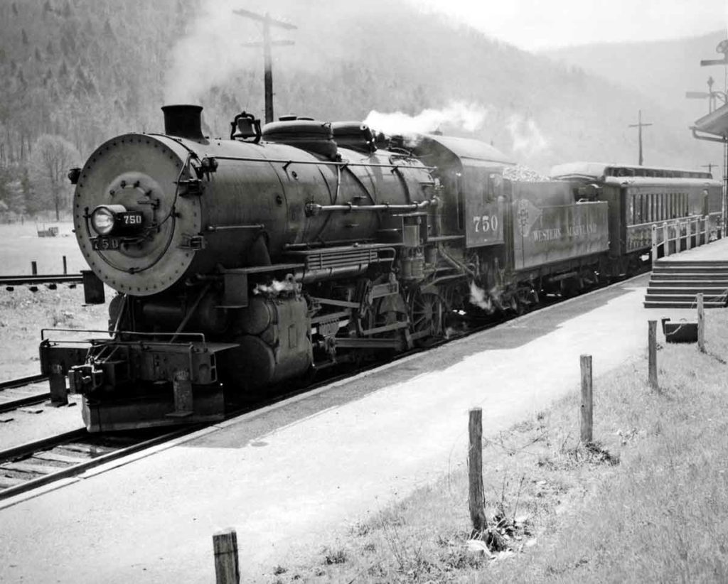 Black and white photo of a train at a station surrounded by mountains.