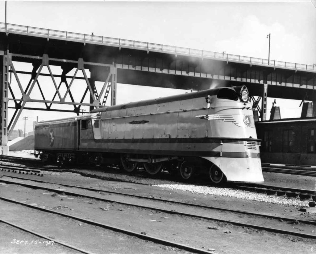 Streamlined 4-4-2 Atlantic-type steam locomotive below bridge