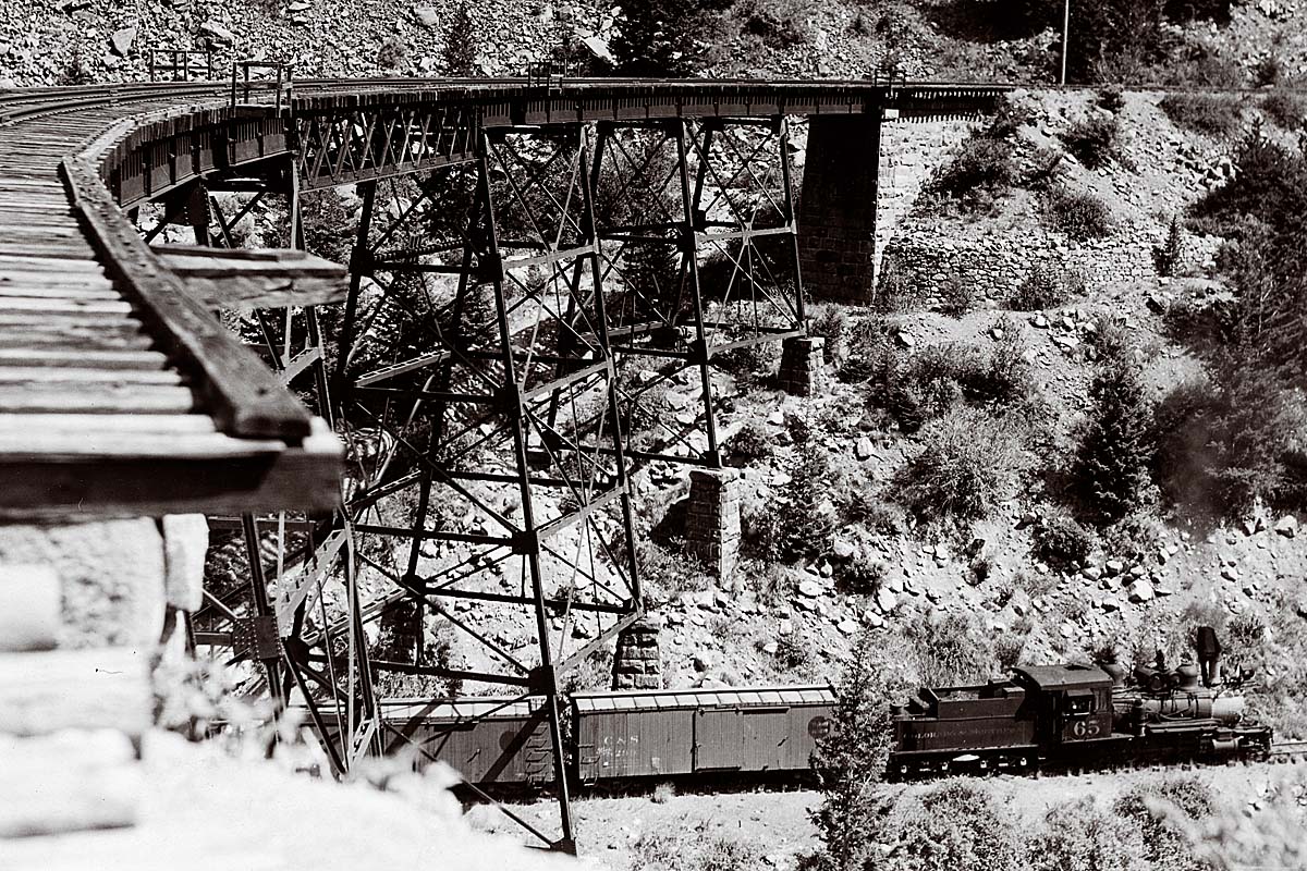 Black and white photo of steam-powered, narrow-gauge freight train crossing over bridge.