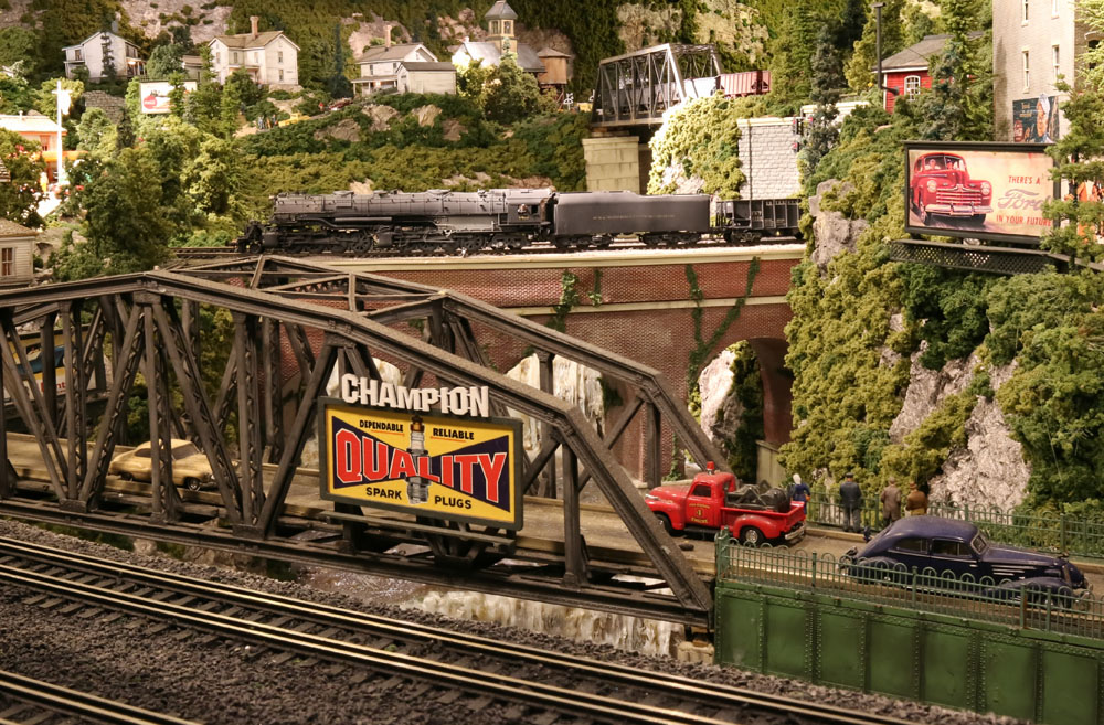 A model 2-8-8-4 steam locomotive pulls a train over a brick viaduct in the background of a verdant city scene.