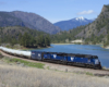 A blue train turning a corner by a body of water with mountains in the background