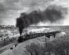 A black and white photo of locomotive Texas & Pacific 2-10-4 with smoke coming out of its chimney. People are standing off to the side of the tracks to let the train pass.
