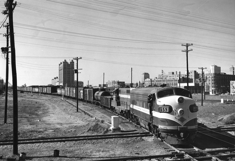A black and white photo of Texas & Pacific F7A 1553 locomotive on the tracks with a city skyline in the background