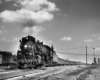 A black and white photo of a brakeman standing on top of locomotive Texas & Pacific 2-8-2 805 as it pulls out of the rail yard