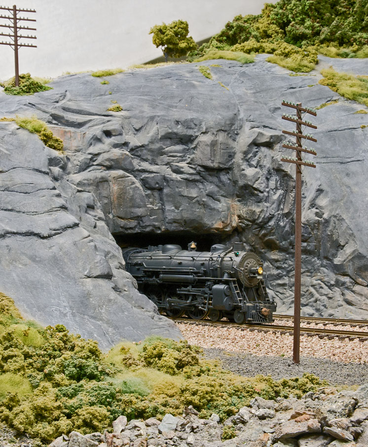 A steam locomotive pokes out of a rock faced tunnel.