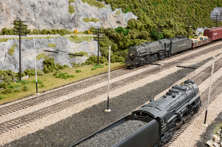Two model trains hauled by steam locomotives passing in a rocky, verdant scene.