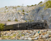 Steam locomotive hauled model express train exits a tunnel in a rocky, mountainous scene.