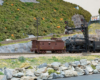 A steam locomotive, tender and caboose pass by signals and a road in a mountainous, verdant scene.