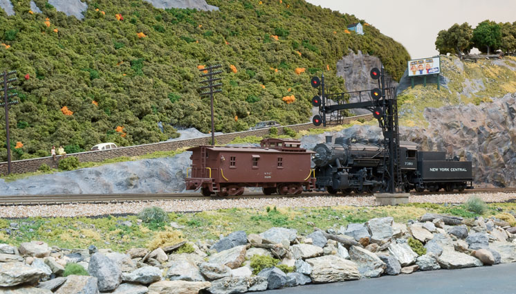 A steam locomotive, tender and caboose pass by signals and a road in a mountainous, verdant scene.