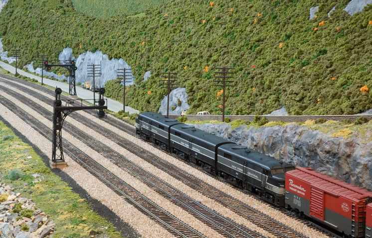Three matching black-and-grey locomotives haul a train on a multi-track mainline in a verdant, mountainous scene.