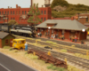 A black and white locomotive pulls a train passed a train station in a brickwork downtown scene.