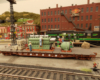 A flatcar with figures and a Diesel engine "load" are stopped in a rail yard amid a brickwork downtown scene.