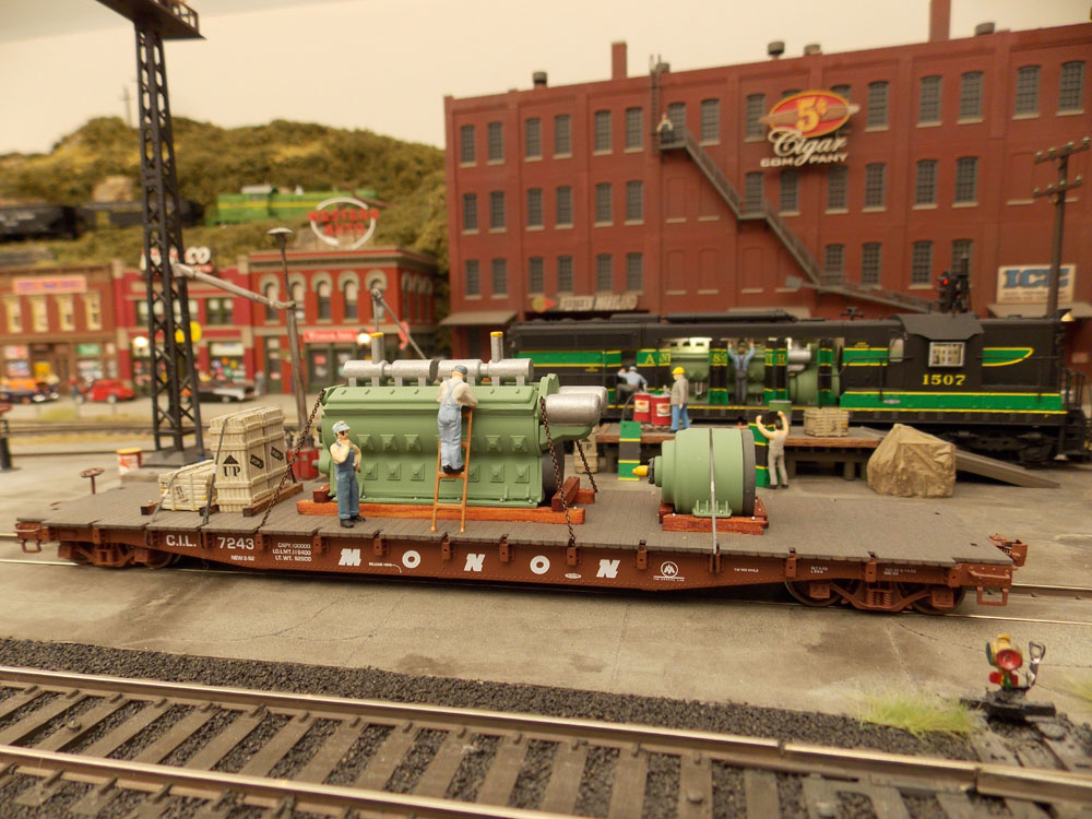 A flatcar with figures and a Diesel engine "load" are stopped in a rail yard amid a brickwork downtown scene.
