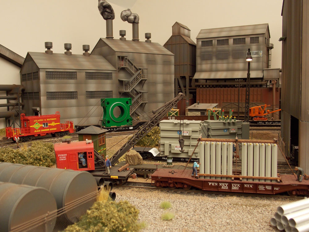 An industrial scene shows a freight train with oversized loads in the background while a crew loads heavy electric equipment on to a flatcar in the foreground.