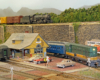 A diesel locomotive and a passenger car near a small town train station. A steam locomotive hauls a freight train on a higher level in the background.