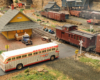 A passenger bus waits at a crossing for a caboose-tailed freight train next to a small town passenger station.