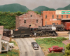 A 2-8-0 steam locomotive hauls a freight train through an industrial scene bracketed by 19th century brick buildings.