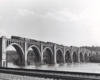 A black and white distant shot of a steam locomotive crossing a bridge