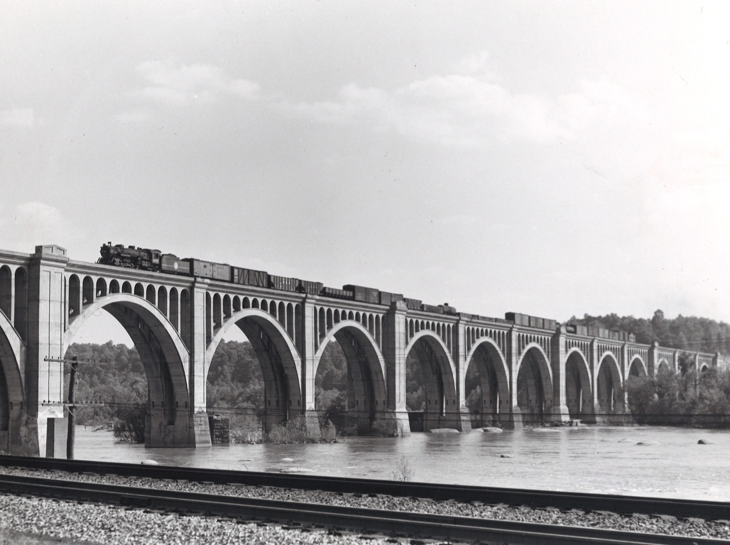 A black and white distant shot of a steam locomotive crossing a bridge