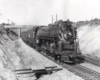 A black and white close up photo of a steam locomotive moving between two hills