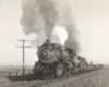 A black and white close up photo of a steam locomotive with white and gray smoke coming out of its chimney