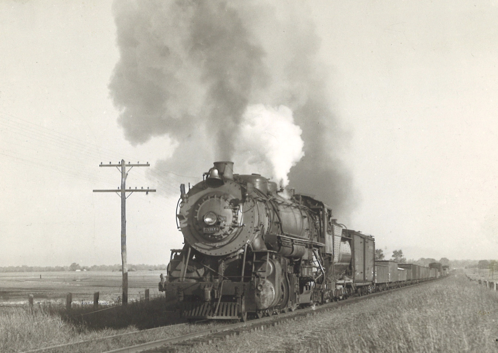 A black and white close up photo of a steam locomotive with white and gray smoke coming out of its chimney