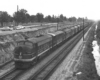 A black and white diesel locomotive on the tracks next to a parking lot with cars in it